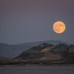 A large orange/yellow moon in a dark sky, above mountains and a body of water.