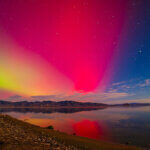 Vibrant rainbow-colored aurora in a starry sky, with mountains in the distance, and all reflected in a body of water in the foreground.