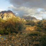 A pebbled stream in the foreground, with tall mountains banded in red in the background. Clouds in a pale blue sky.