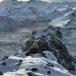 A snowy mountain range with snowy roads below. Cloudy skies.