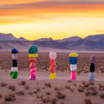 An art installation in the desert, with large stacked boulders painted in vibrant colors. Mountains and an orange/yellow sunset in the background.