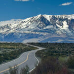 A paved road winds its way through the desert, toward snowy mountains in the distance. Puffy white clouds in a blue sky.