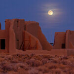 A bright full moon cuts through wispy clouds, behind clay ruins in the desert.