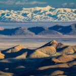 Layered landscape of brown scrub-covered desert mountains, water, and snowy mountains in the background.