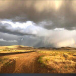 A stormy golden desert scene, with a road disappearing into the distant mountains, and storm clouds dropping rain. There is a slight rainbow in the distance.