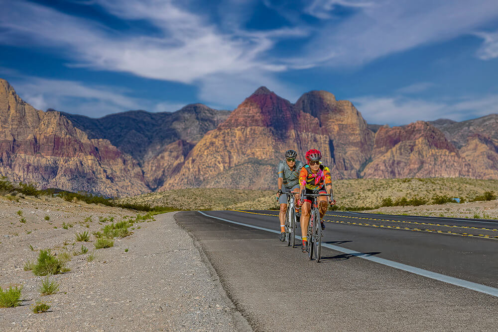 Two people riding bikes along a road that disappears into tall red-ribboned mountains in the background. Soft white clouds in a blue sky.