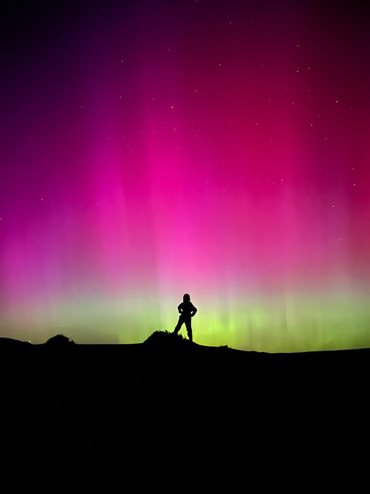A person stands on a hillside, silhouetted in front of strong pink and white striated aurora, with stars showing through.