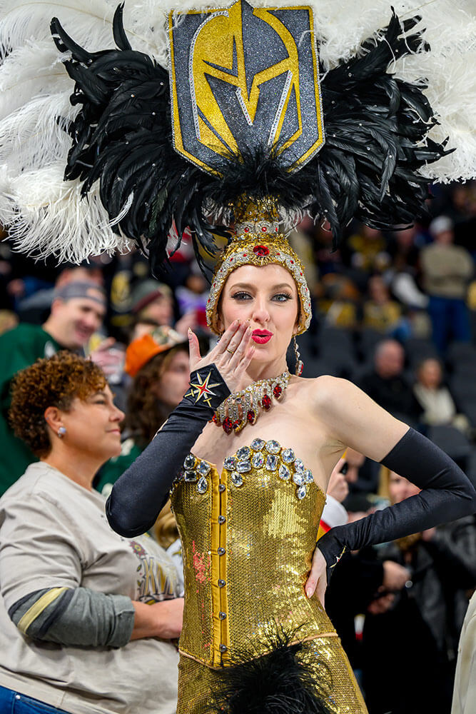 A woman dressed in yellow, gray, and black sequined outfit, with a large feathered headdress, seems to blow a kiss to the photographer. There are crowds of people behind her.