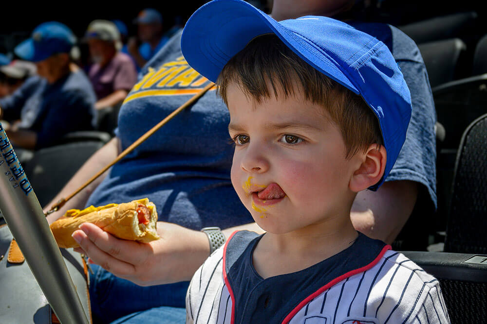 A young baseball enthusiast licks mustard off his lips while an adult holds onto a hot dog behind him.