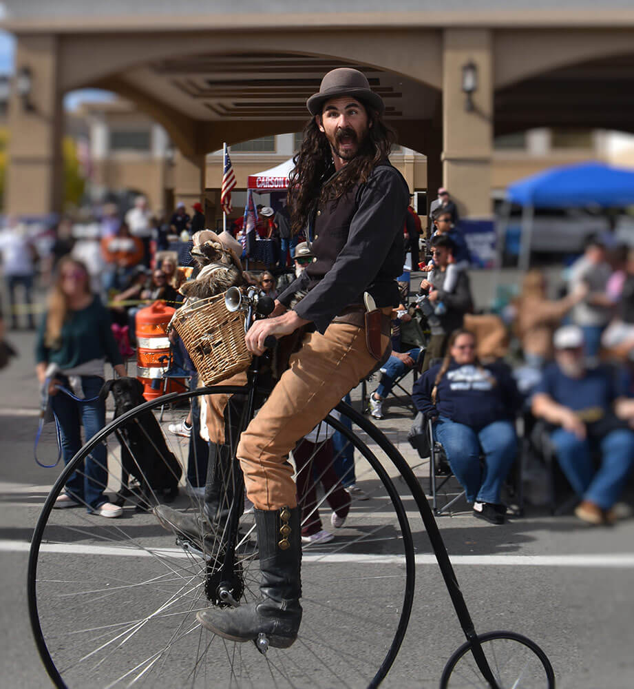 A man rides a penny-farthing, or high-wheel bicycle. There are crowds of people behind him.
