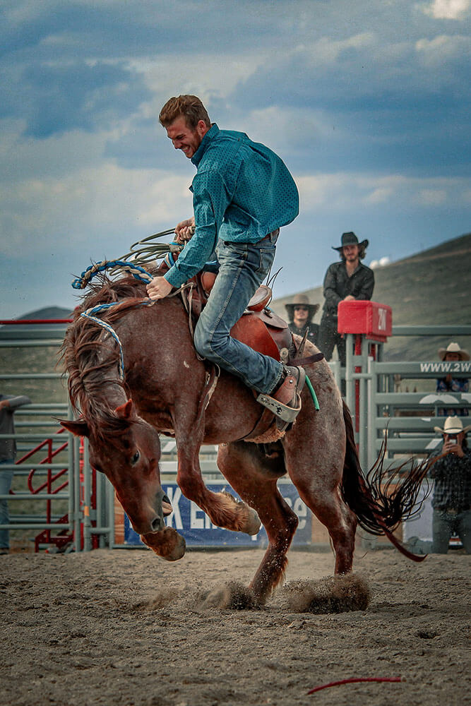 A man rides a bucking horse at a rodeo. Other men in the background, with a cloudy sky.
