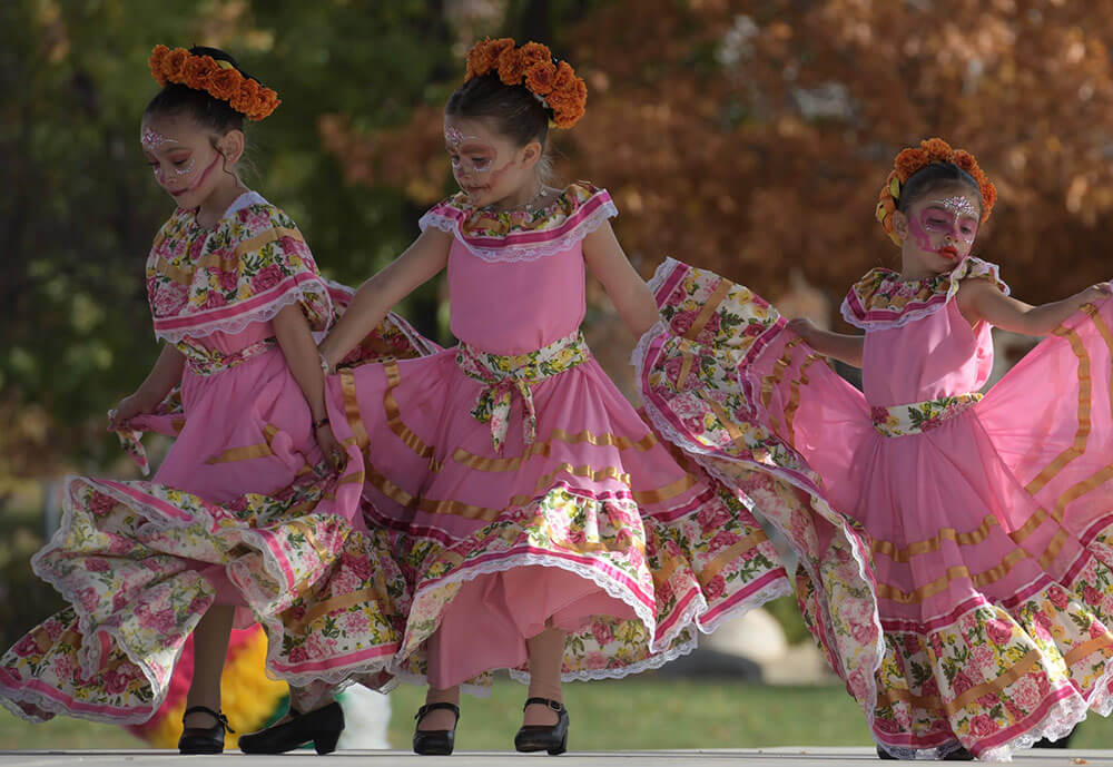 Three young girls in pink flowery dresses and face paint, dance on a stage.