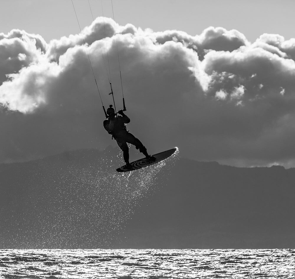 A black and white image of a person whose feet are on a small surfboard that looks like it's just lifted out of the water. The person is holding onto strings that presumably go to some sort of kite. There are mountains and stormy clouds in the background, and the person is mostly silhouetted.