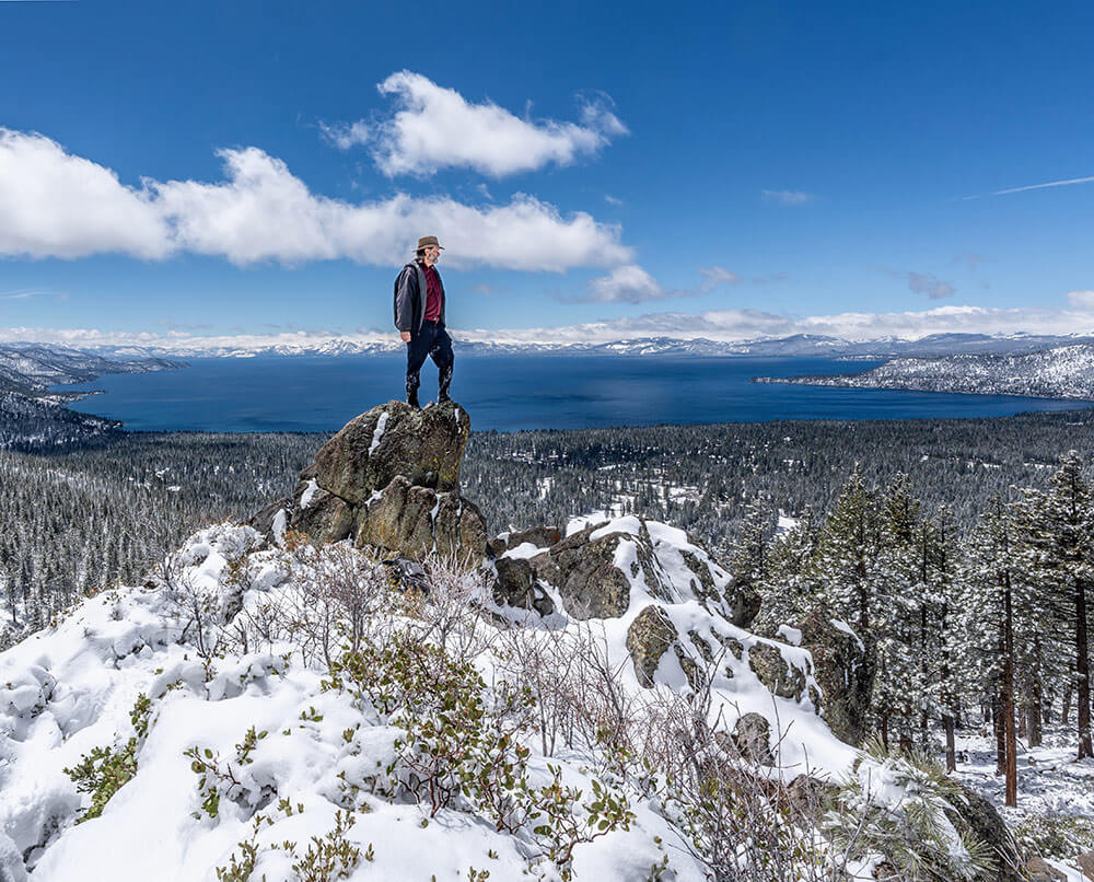 A man stands on top of a snowy rock, overlooking forest and Lake Tahoe in the background, with snowy mountains all around and puffy white clouds in a blue sky.