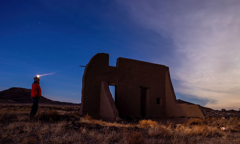 A person wearing a headlamp stands beside ruins of a clay building. Partly cloudy sky, with stars showing through.