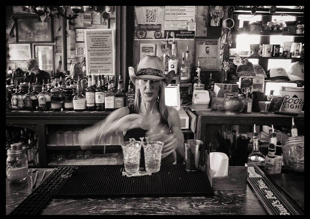 Black and white image of a woman bartender wearing a cowboy hat and mixing a drink.