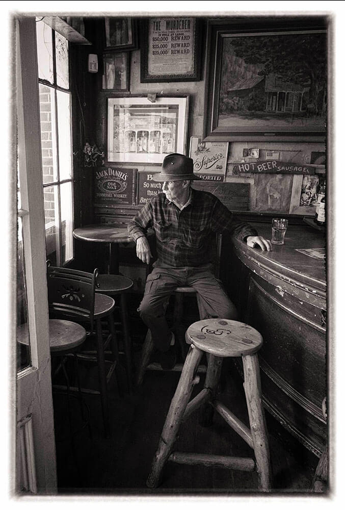 Black and white image of a man sitting on a barstool in what looks like an old bar, looking out a window.