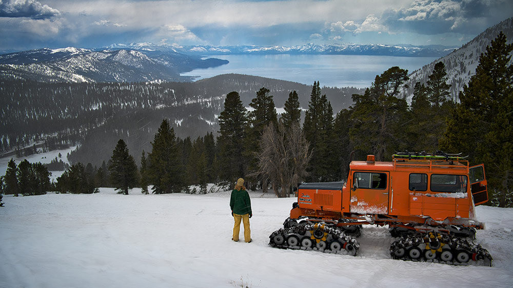 A person stands on a snowy hillside, with a heavy-duty snow vehicle, overlooking forests and Lake Tahoe, with snowy mountains surrounding.