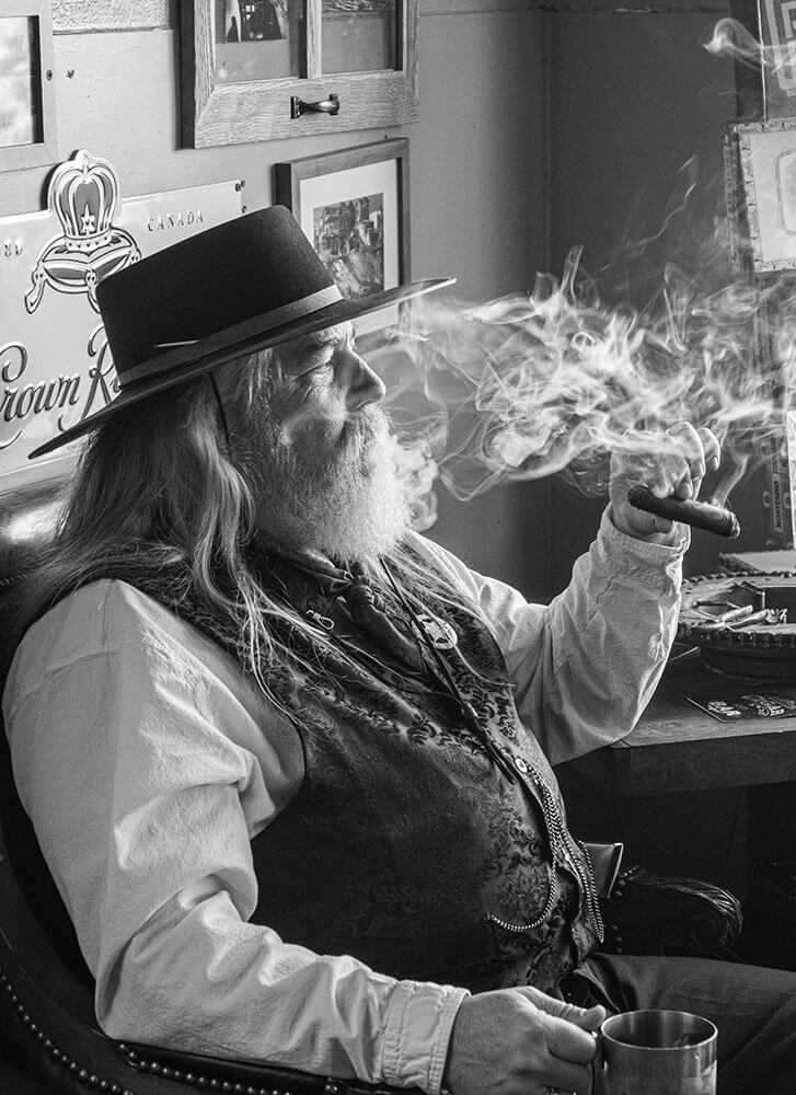 Black and white photo of a person dressed in old-time clothing, smoking a cigar, with backlit cigar smoke swirling around.