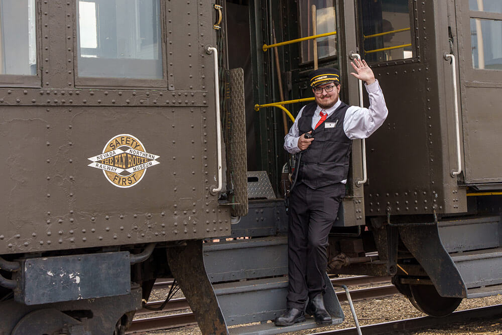 A train conductor stands on a step of a train and waves at the camera.