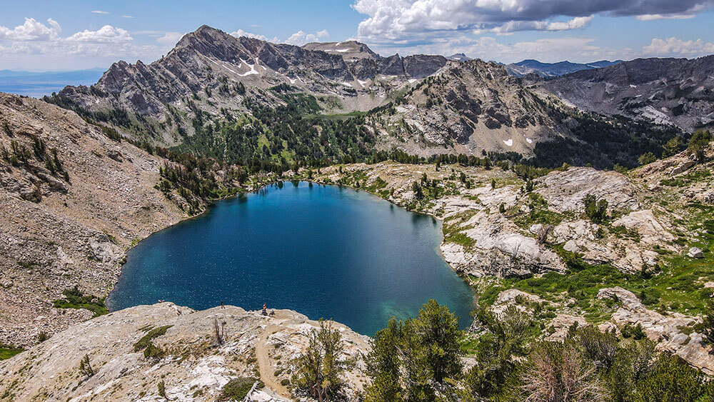 Aerial shot of a remote mountain lake, surrounded by trees, mountains and desert plants. Clouds in a blue sky.