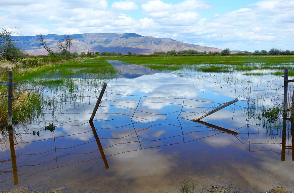 A flooded field with sagging wire fencing. Mountain and cloudy sky are reflected in the still water.