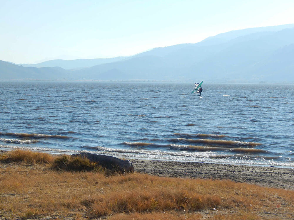 A person windsurfing out in a mountain lake. Mountains in the background, light blue sky.