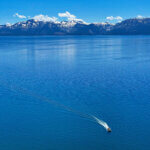 An aerial shot of a motorboat on Lake Tahoe, with snowy mountains and cloudy blue skies in the background.