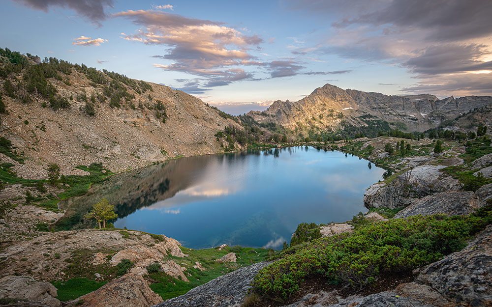A remote mountain lake, with trees, mountains, and clouds in a blue sky reflected in the water.