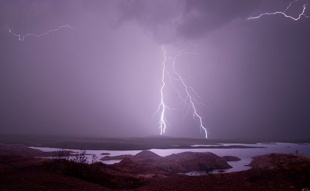 Purple-tinted image of a desert lake with lightning strikes in a storm.