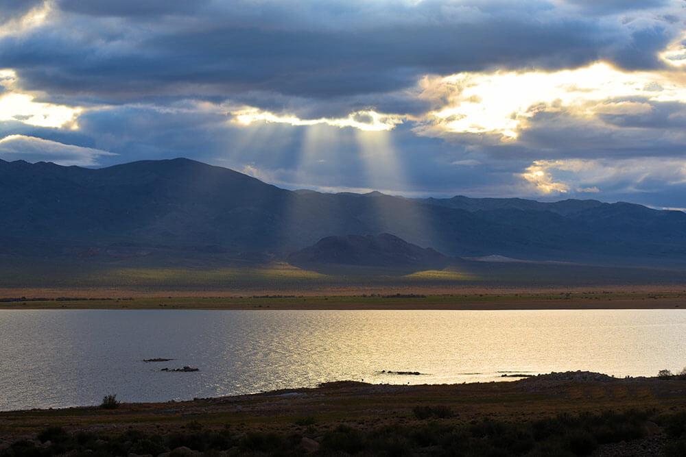 Desert lake with mountains in the background and crepuscular rays of sunlight breaking through a cloudy sky.
