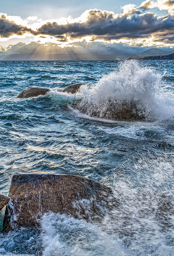 Stormy water crashing over rocks, with mountains in the background and crepuscular rays of sunlight breaking through the clouds in the sky.