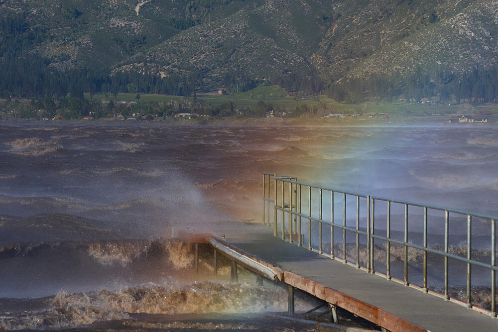 Stormy desert lake with a walkway leading out to it and a rainbow caused by the water spray. Green mountains in the background.