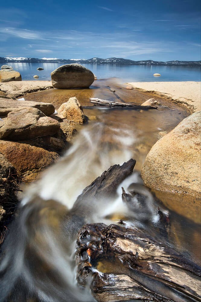 A long-exposure photo of a river coming off a large lake, with snowy mountains and streaky blue skies in the background.