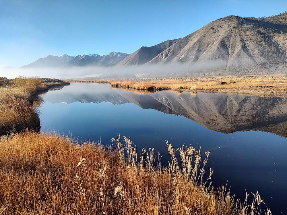 Desert river, with low-hanging clouds along the base of tall mountains annd blue skies in the background, all reflected in the water.