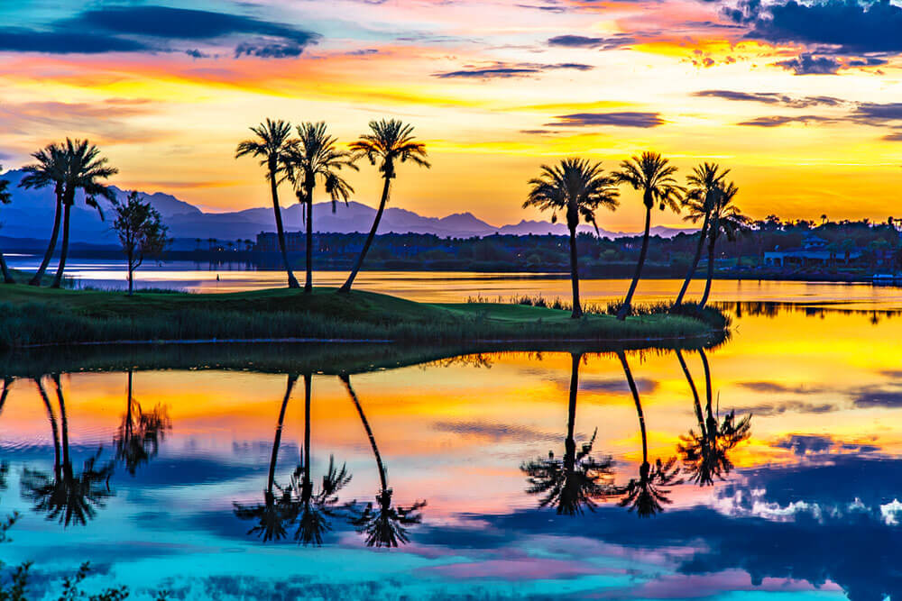 Desert lake with mountains in the background, an a small peninsula with palm trees silhouetted against a yellow and orange sunset, all reflected in the water.